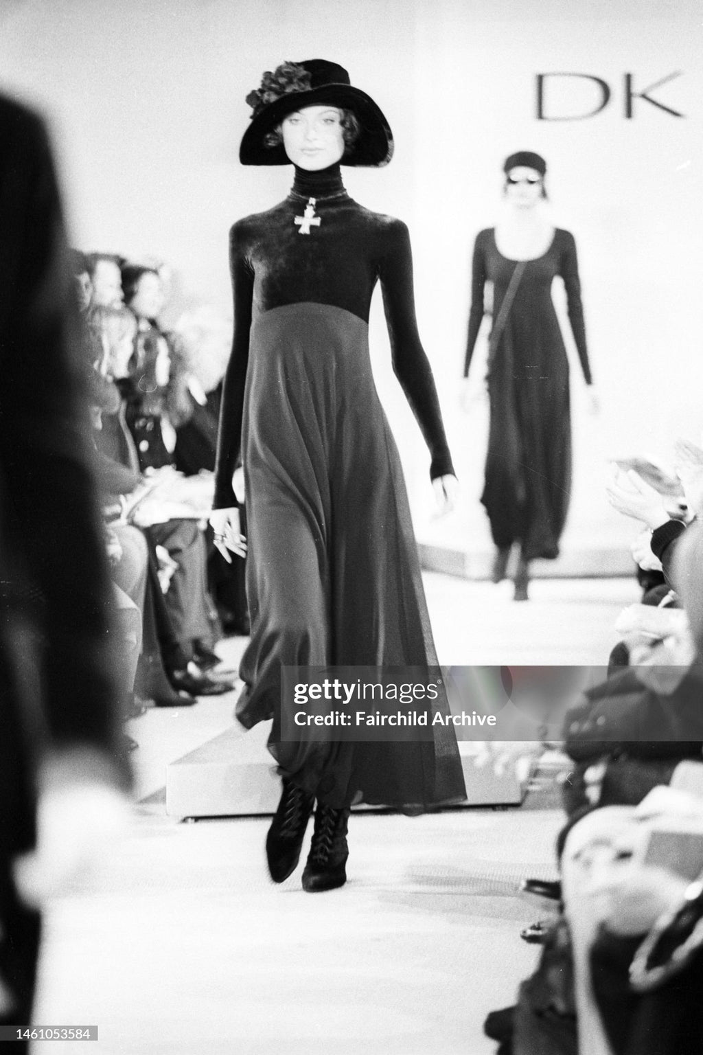 Black and white runway photo showing a model walking down a fashion show catwalk in a long dark dress and wide-brimmed hat, with a DK logo visible in the background.